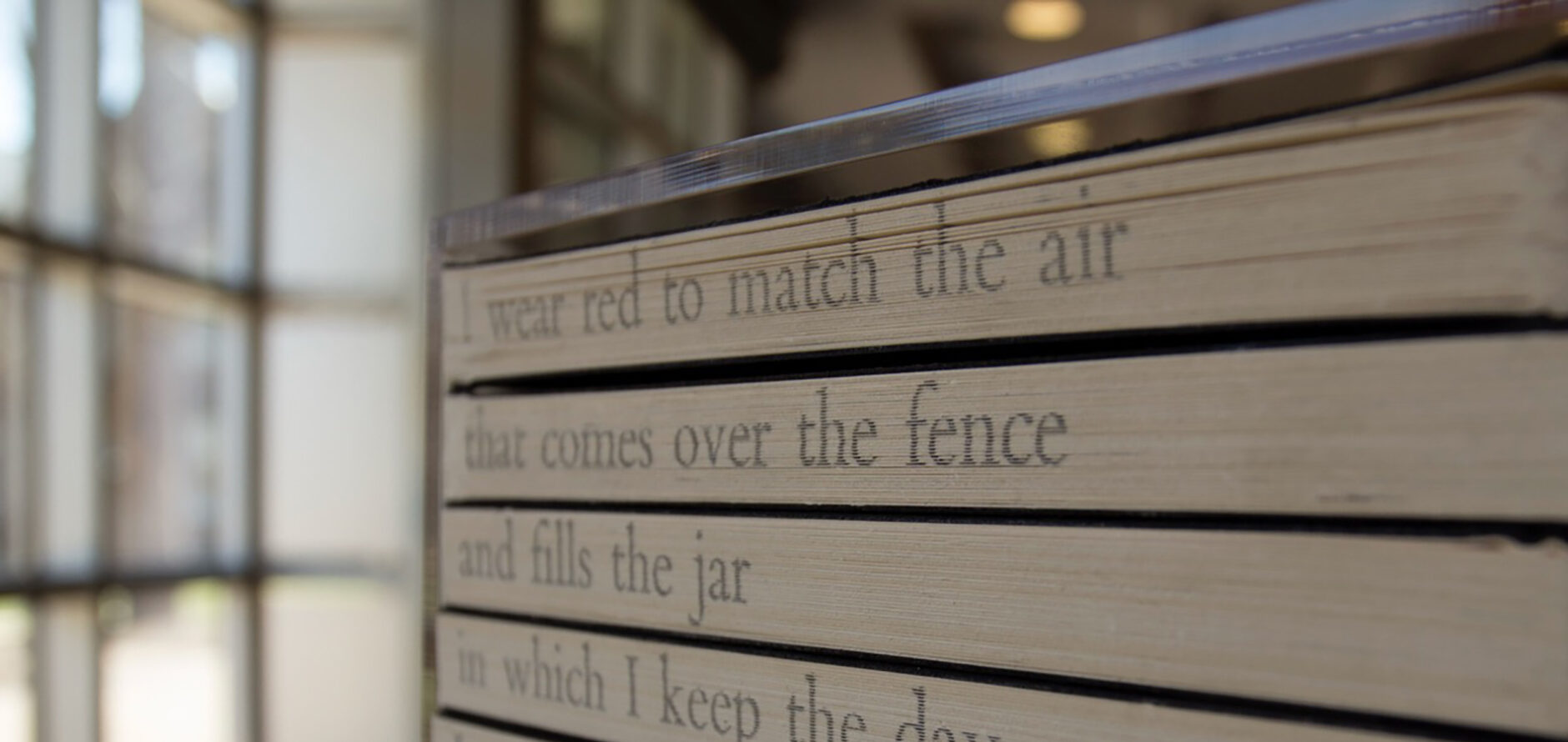 A sculpture of spine-in books on a shelf with the beginnings of a poem written on the pages that reads: I wear red to match the air / that comes over the fence / and fills the jar / in which I keep the day...