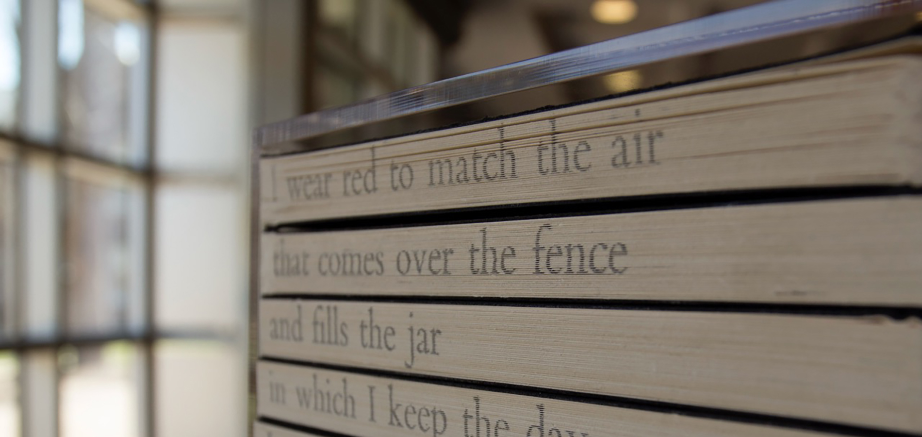A sculpture of spine-in books on a shelf with the beginnings of a poem written on the pages that reads: I wear red to match the air / that comes over the fence / and fills the jar / in which I keep the day...