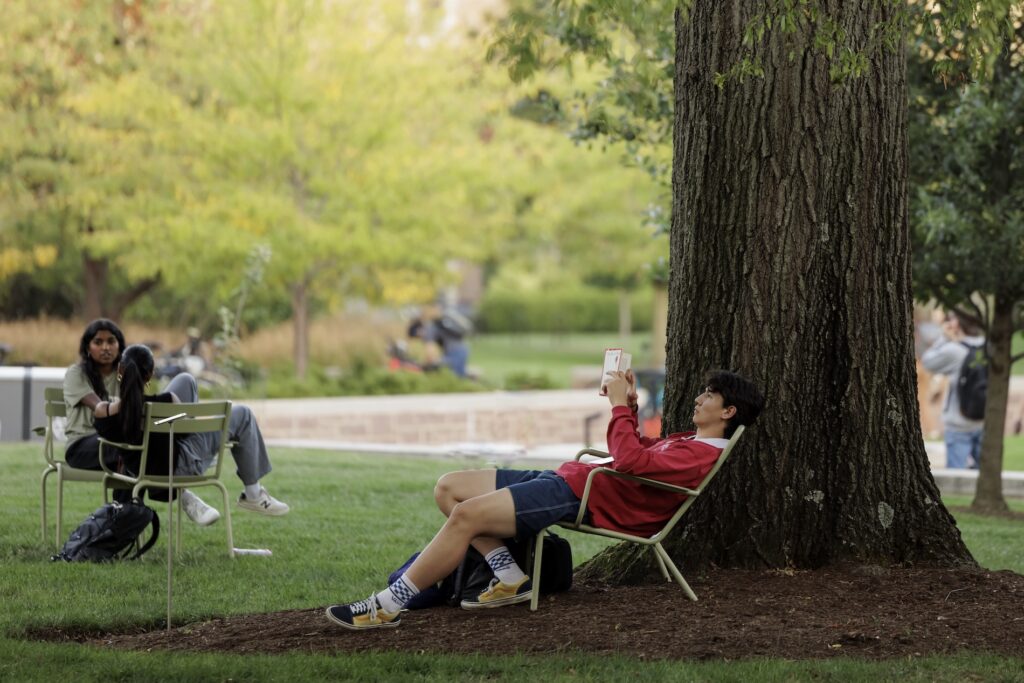 A student reads outside under a tree as other students talk.