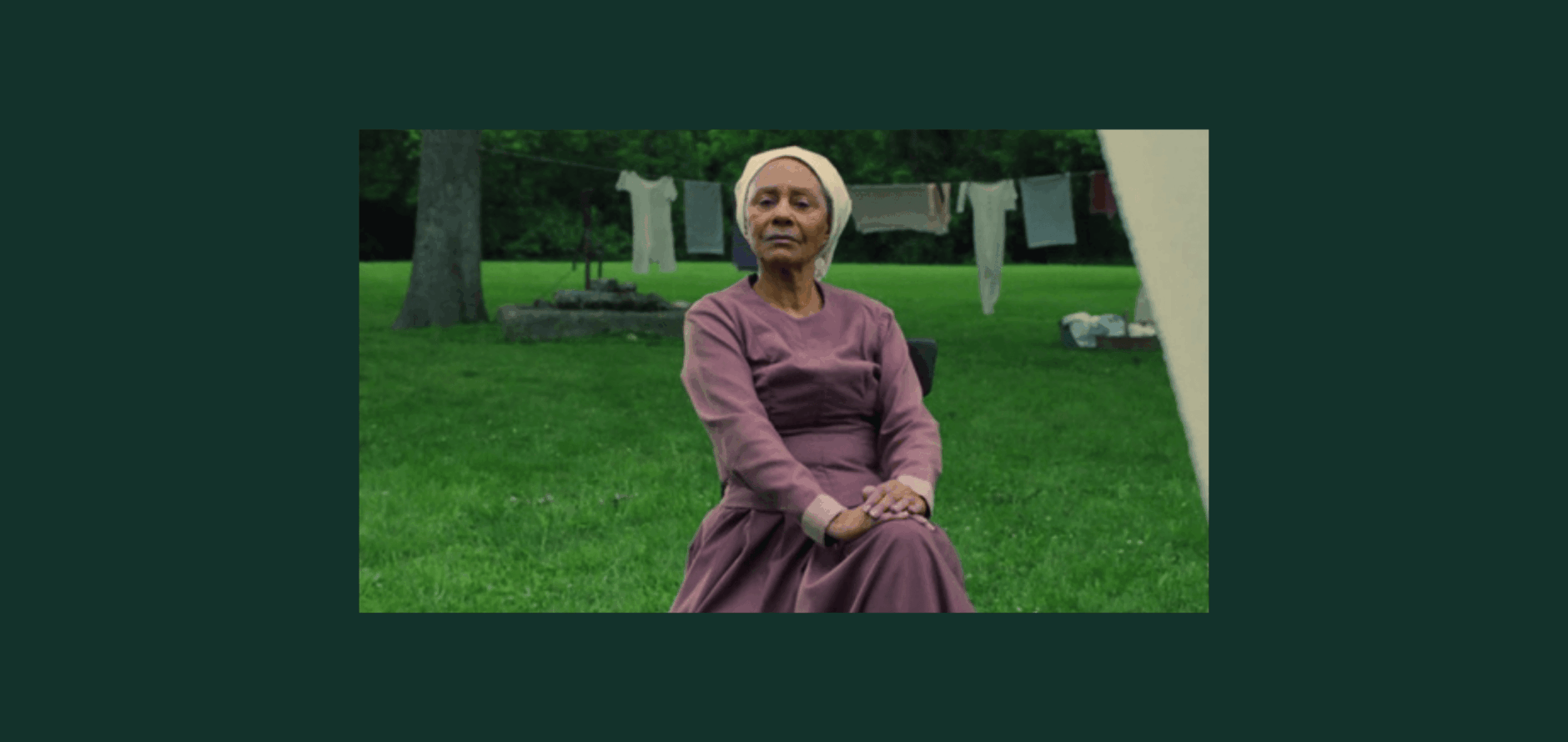 Image from the short film PELAGIE X. In the image, an older Black woman wearing a mauve long-sleeved dress and a light headscarf sits outdoors on a green lawn with her hands folded in her lap. Behind her, laundry hangs on clotheslines between trees.