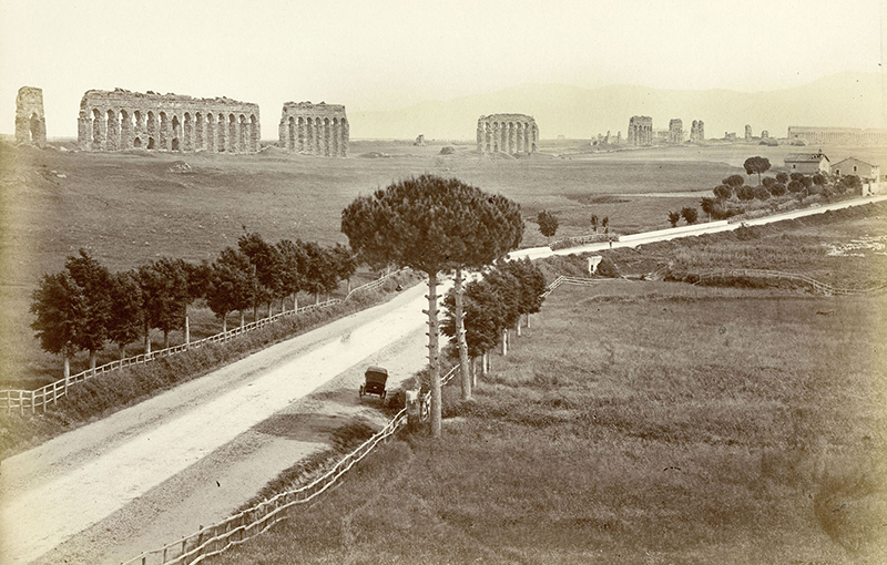 Photograph of Aventine Hill in Rome circa May 1925. A dirt road bracketed by a wooden fence and equally spaced trees is featured. A buggy is pulled along the side of the road in the foreground and the background features arched ruins.