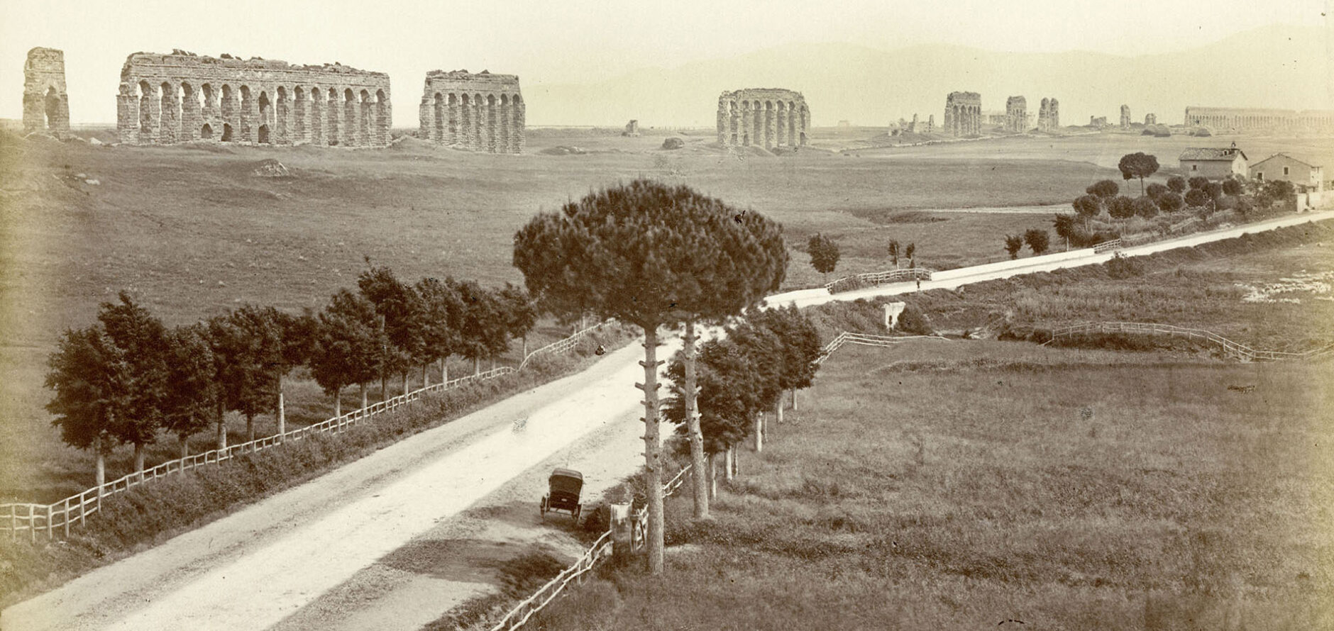 Photograph of Aventine Hill in Rome circa May 1925. A dirt road bracketed by a wooden fence and equally spaced trees is featured. A buggy is pulled along the side of the road in the foreground and the background features arched ruins.