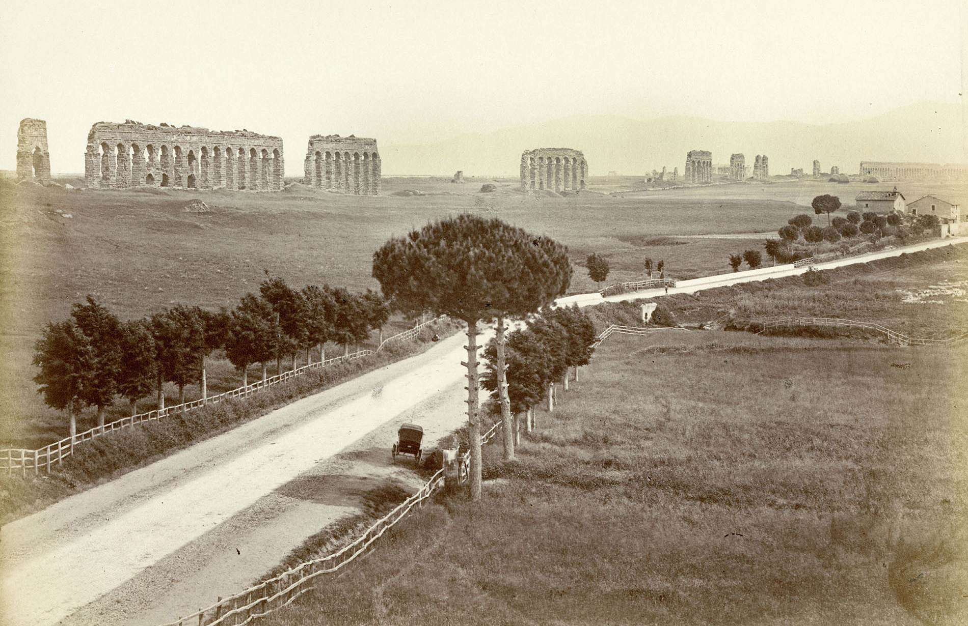 Photograph of Aventine Hill in Rome circa May 1925. A dirt road bracketed by a wooden fence and equally spaced trees is featured. A buggy is pulled along the side of the road in the foreground and the background features arched ruins.