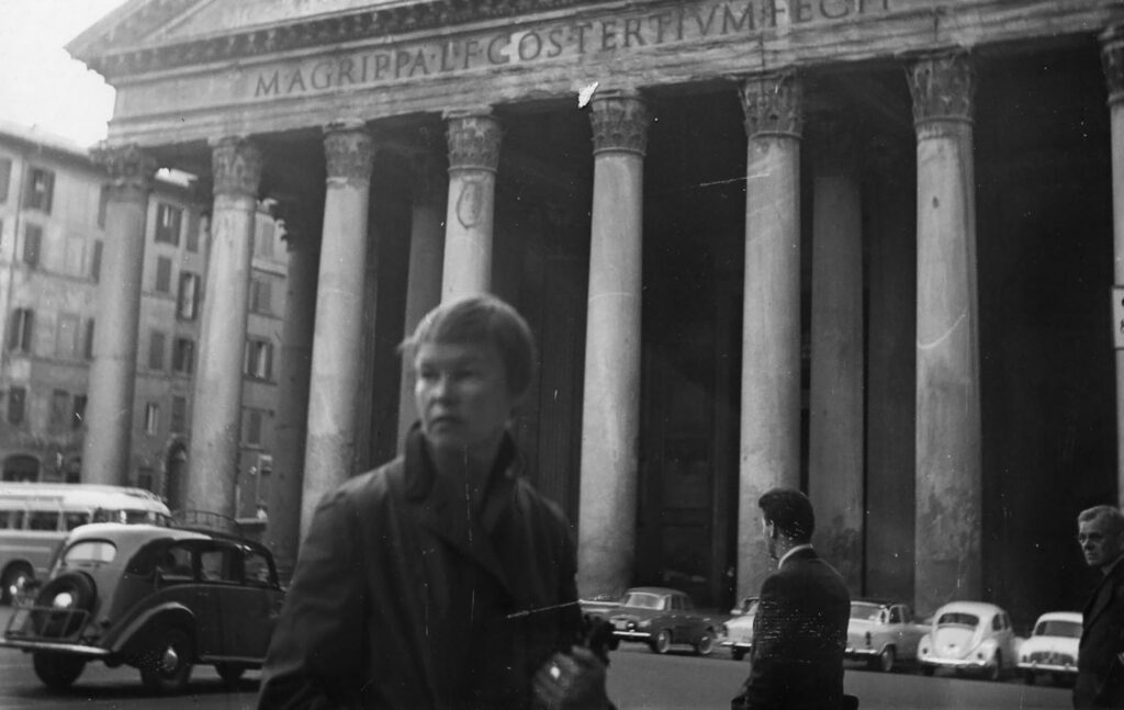 May Swenson in a coat clutching her purse in front of the Pantheon.