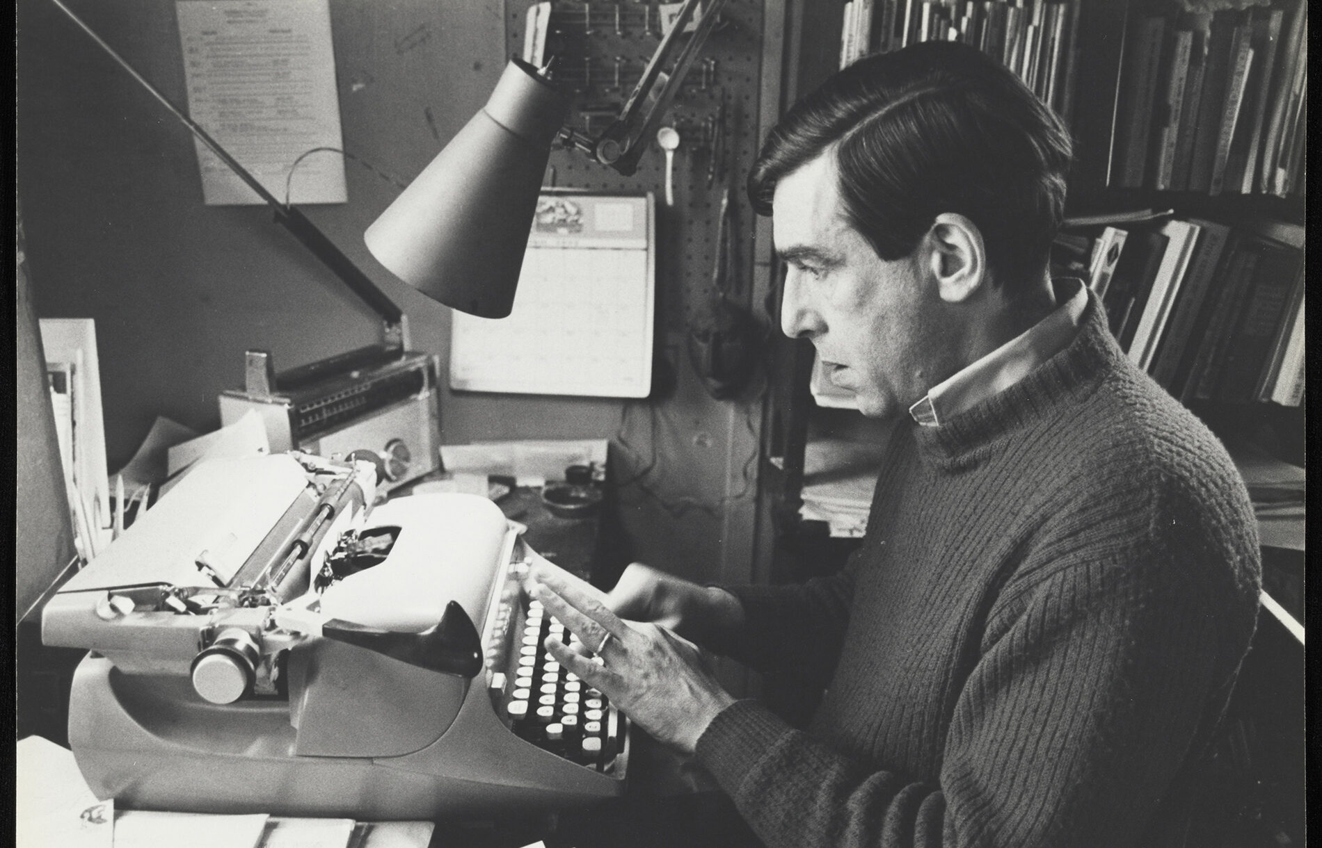 Russell Edson sat typing at a paper-strewn desk in front of a typewriter in a crowded little office space.