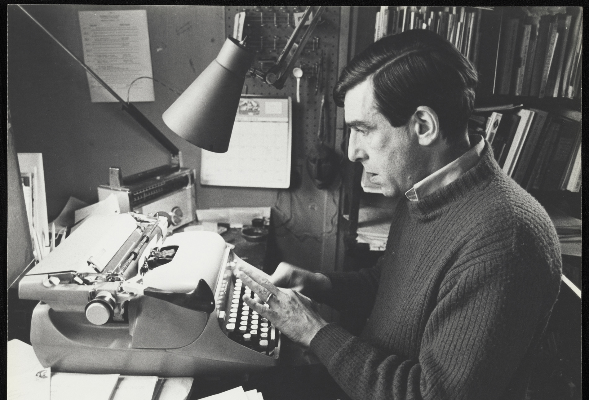 Russell Edson sat typing at a paper-strewn desk in front of a typewriter in a crowded little office space.
