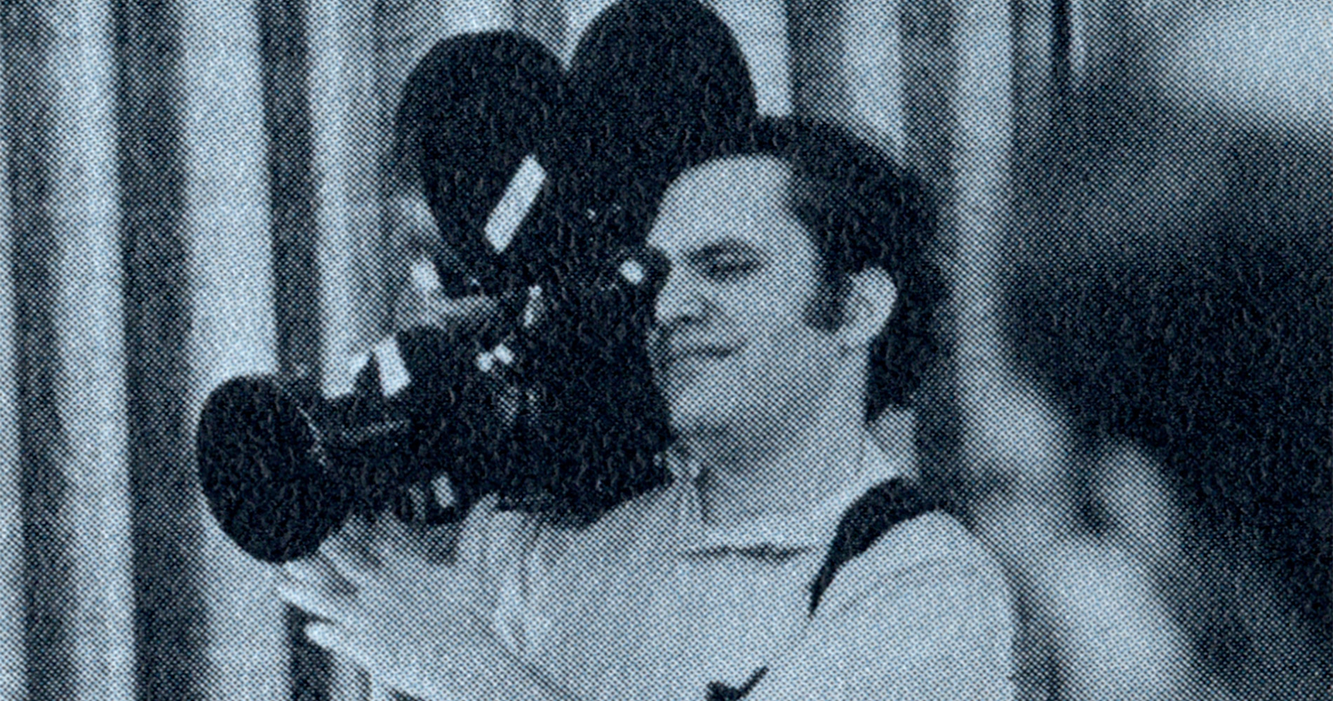 A man standing with a film camera on his shoulder and his eye on the view-piece.