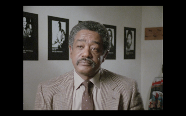 A Black man with a moustache sits for an interview in a suit and tie.
