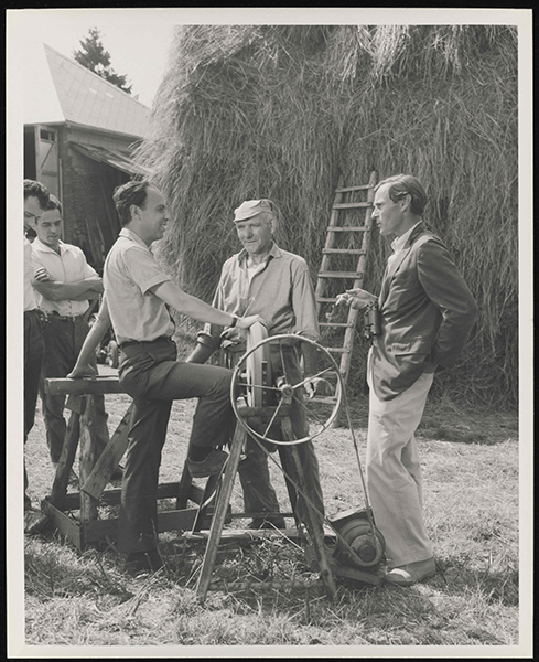 A man in a suit jacket and slacks stands speaking with another man in a short sleeved button up while three other men in collared button-ups listen in the background.