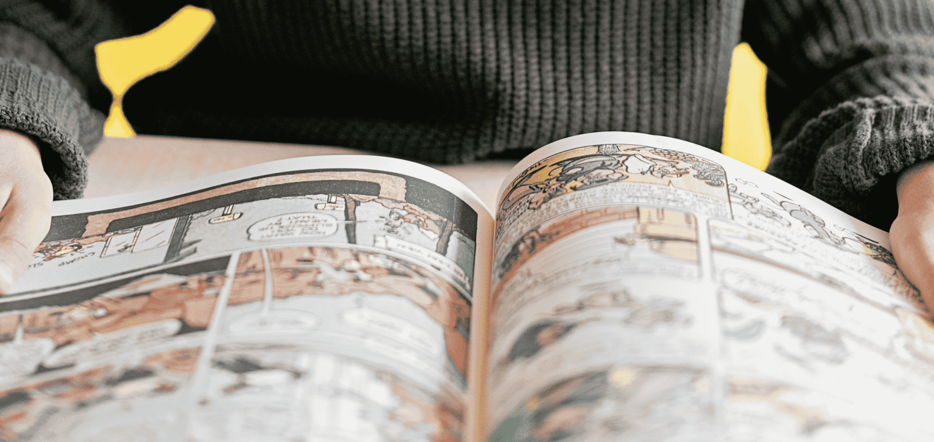A person in a dark sweater with yellow accents holds open a graphic novel, showing illustrated comic panels across both pages.