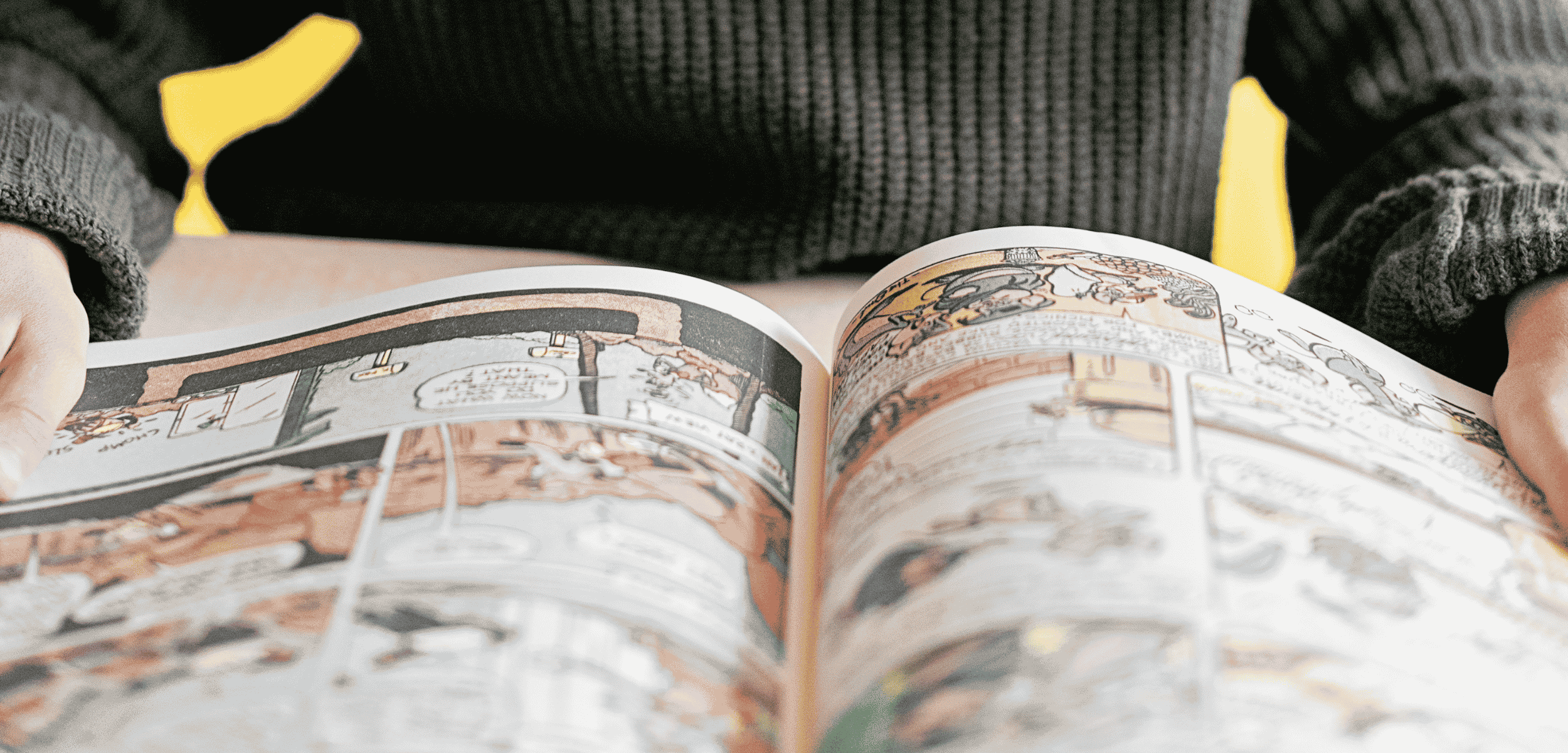 A person in a dark sweater with yellow accents holds open a graphic novel, showing illustrated comic panels across both pages.
